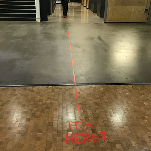 The words  It's here in red taped on the floor with a red line leading out of frame